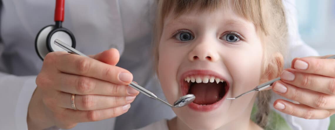 Portrait of joyful little girl visiting dentist and looking at camera with joyfulness. Doctor in white medical gown check-up oral cavity of baby. Medicine and healthcare concept