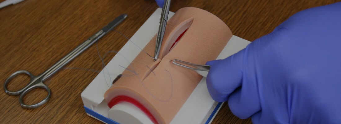 a man holding forceps and scalpel and practicing on regular suture pad with scissor around